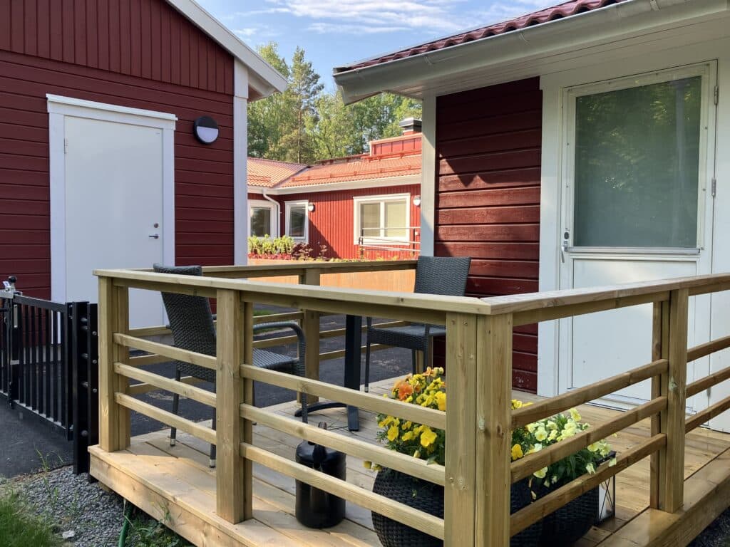 Veranda utanför rött hus med vita knutar. På verandan står utemöbler och en kruka med gula blommor.