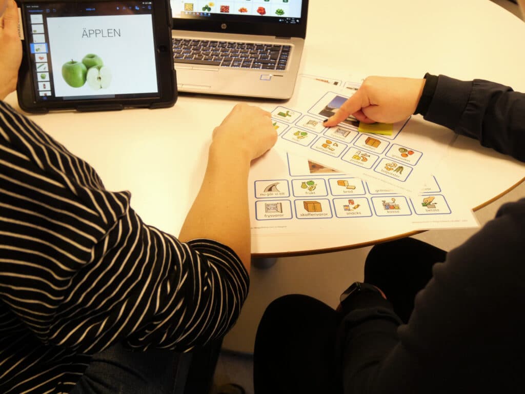 Två personer som sitter vi ett bord och pekar på bildstöd med en Ipad brevdid sig där det står "äpplen" på skärmen.