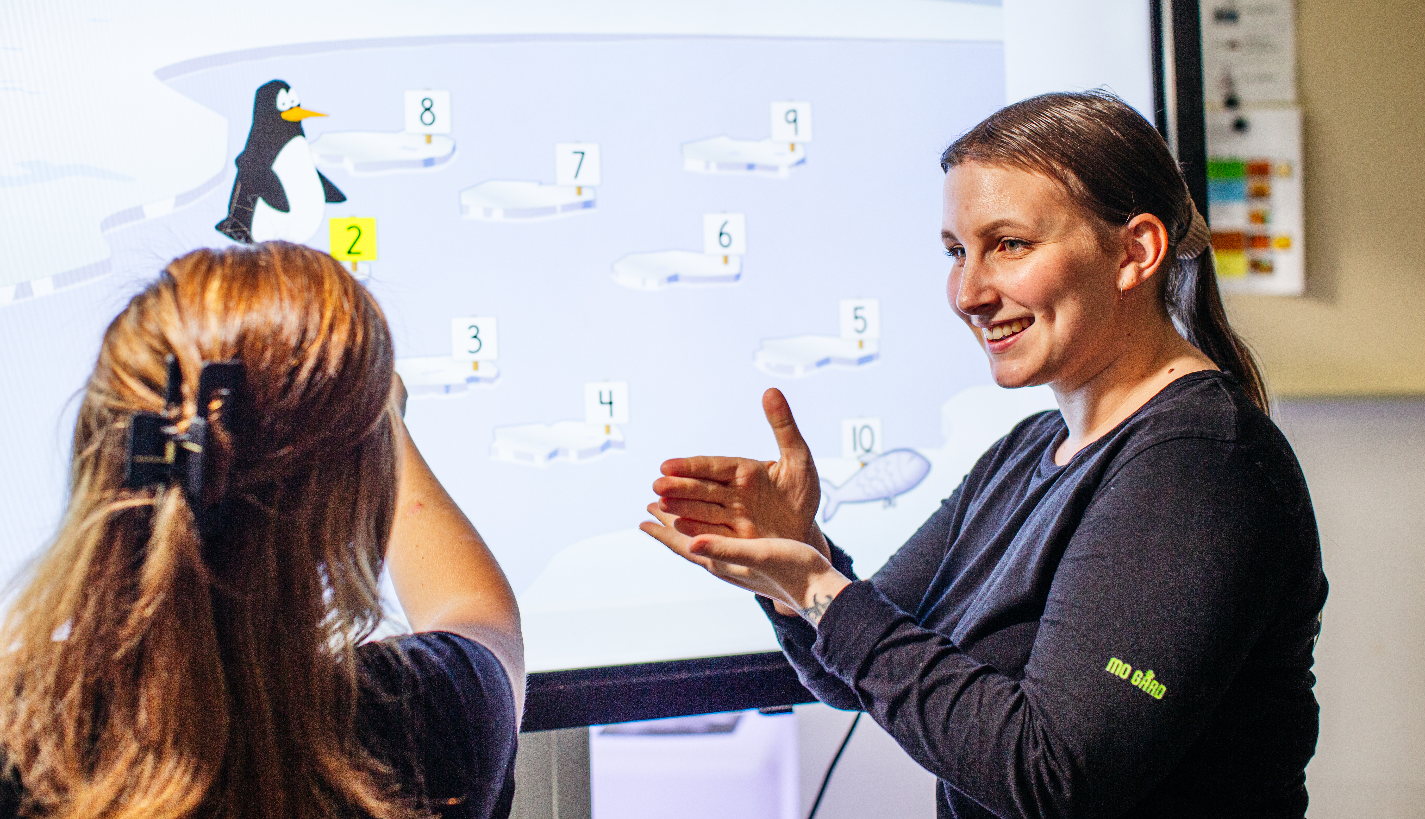 En lärare och en elev står framför en interaktiv skärm som visar ett spel med en tecknad pingvin och numrerade isflak. Läraren ler och gestikulerar med händerna medan eleven pekar mot skärmen med ryggen vänd mot kameran.