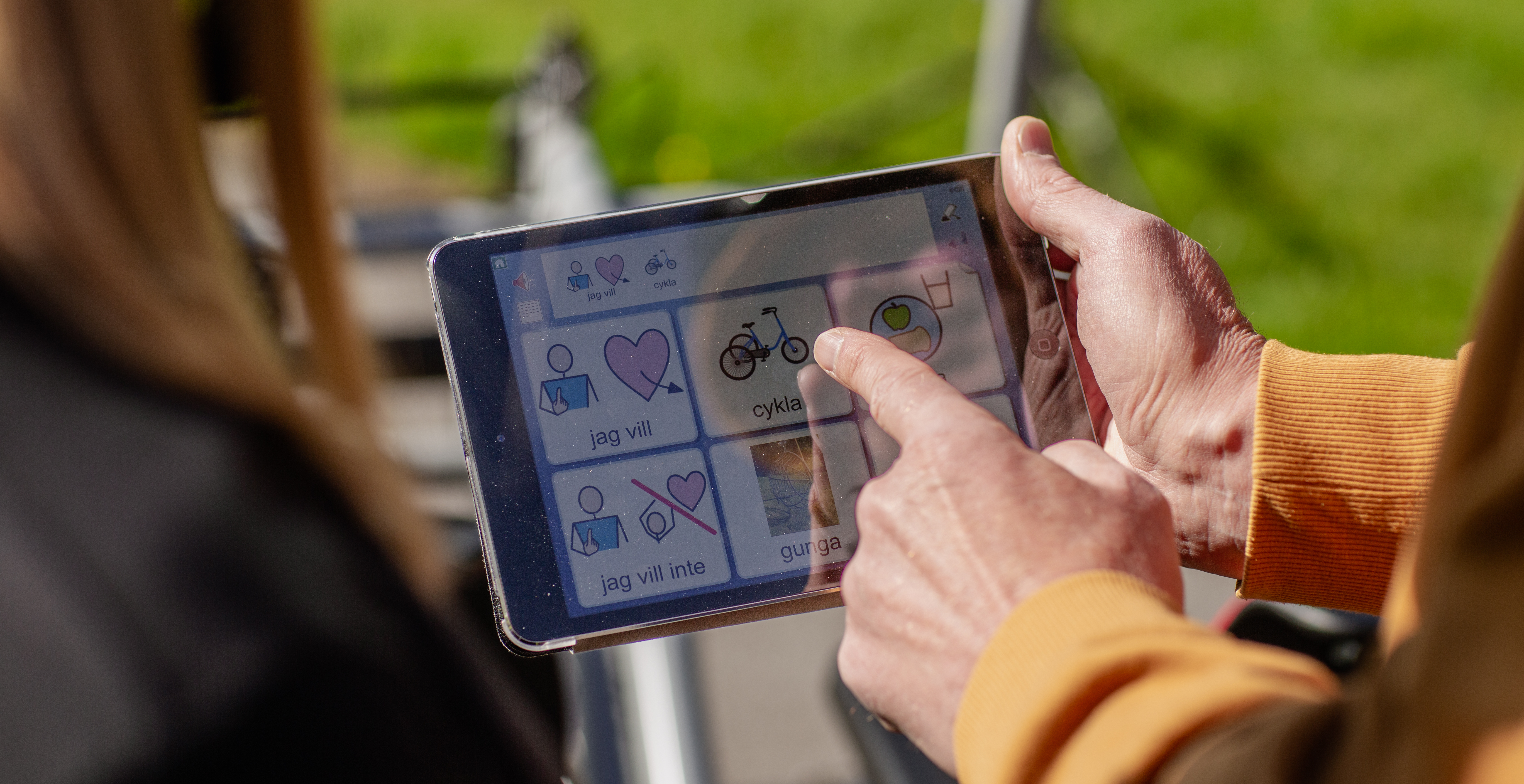 En person använder en surfplatta med bildstöd för kommunikation. På skärmen syns symboler med text som ”jag vill”, ”cykla” och ”jag vill inte”. Personen pekar på symbolen för cykla.