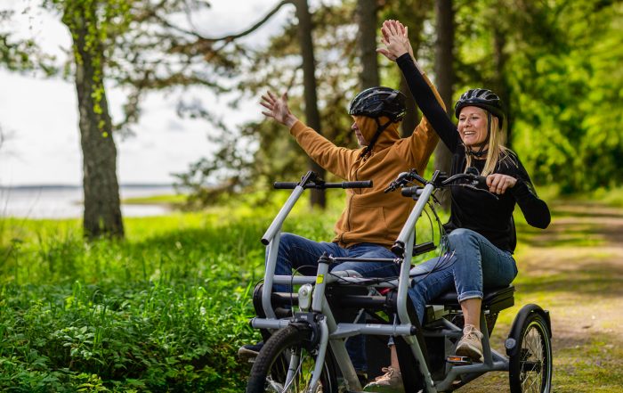 Två personer med cykelhjälmar sitter tillsammans på en trehjulig cykel på en skogsstig. De håller upp händerna och ser glada ut medan de cyklar genom det gröna landskapet.