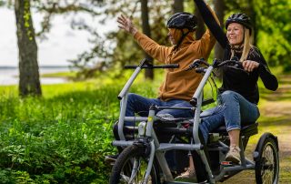 Två personer med cykelhjälmar sitter tillsammans på en trehjulig cykel på en skogsstig. De håller upp händerna och ser glada ut medan de cyklar genom det gröna landskapet.