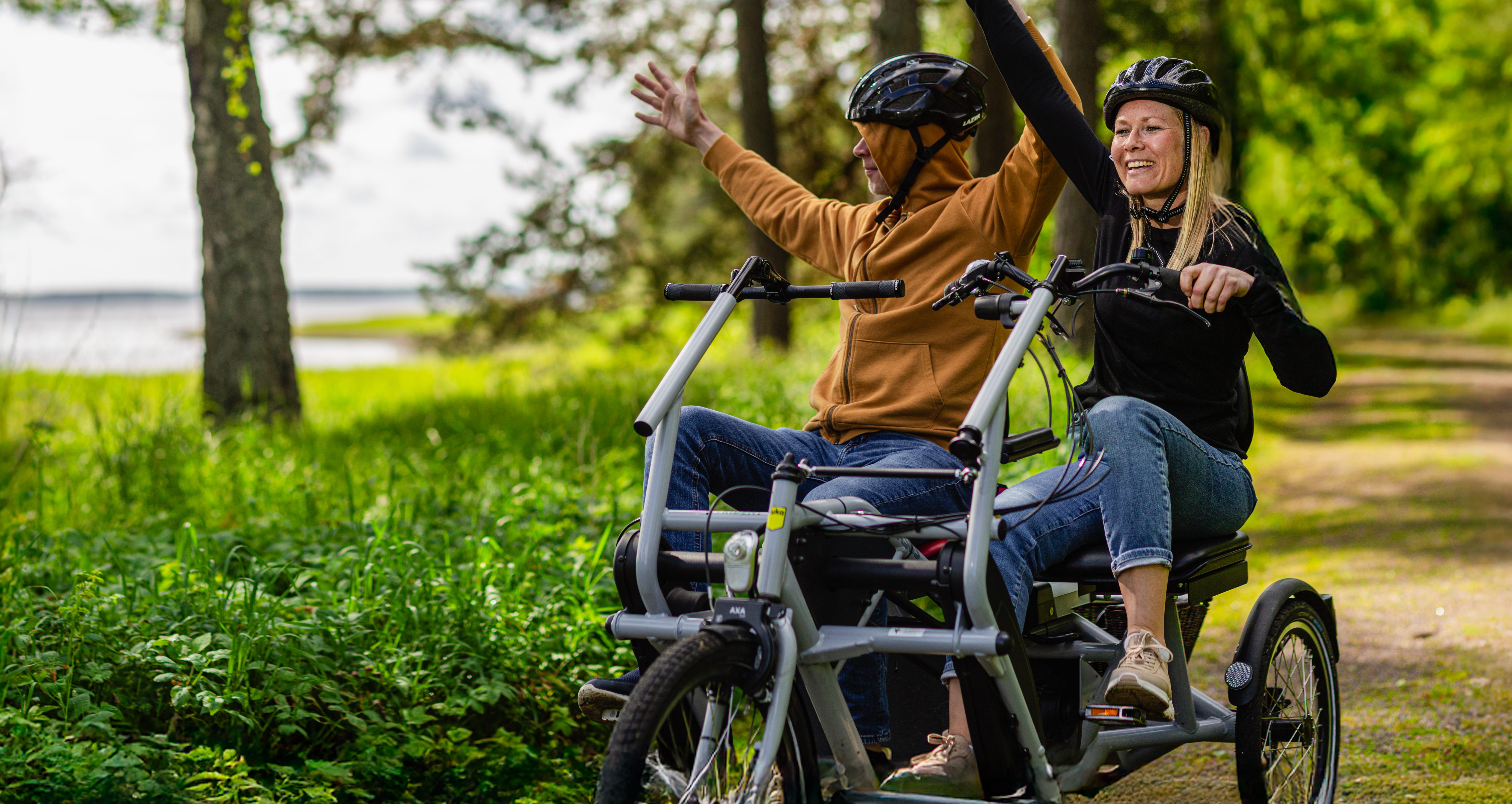 Två personer med cykelhjälmar sitter tillsammans på en trehjulig cykel på en skogsstig. De håller upp händerna och ser glada ut medan de cyklar genom det gröna landskapet.