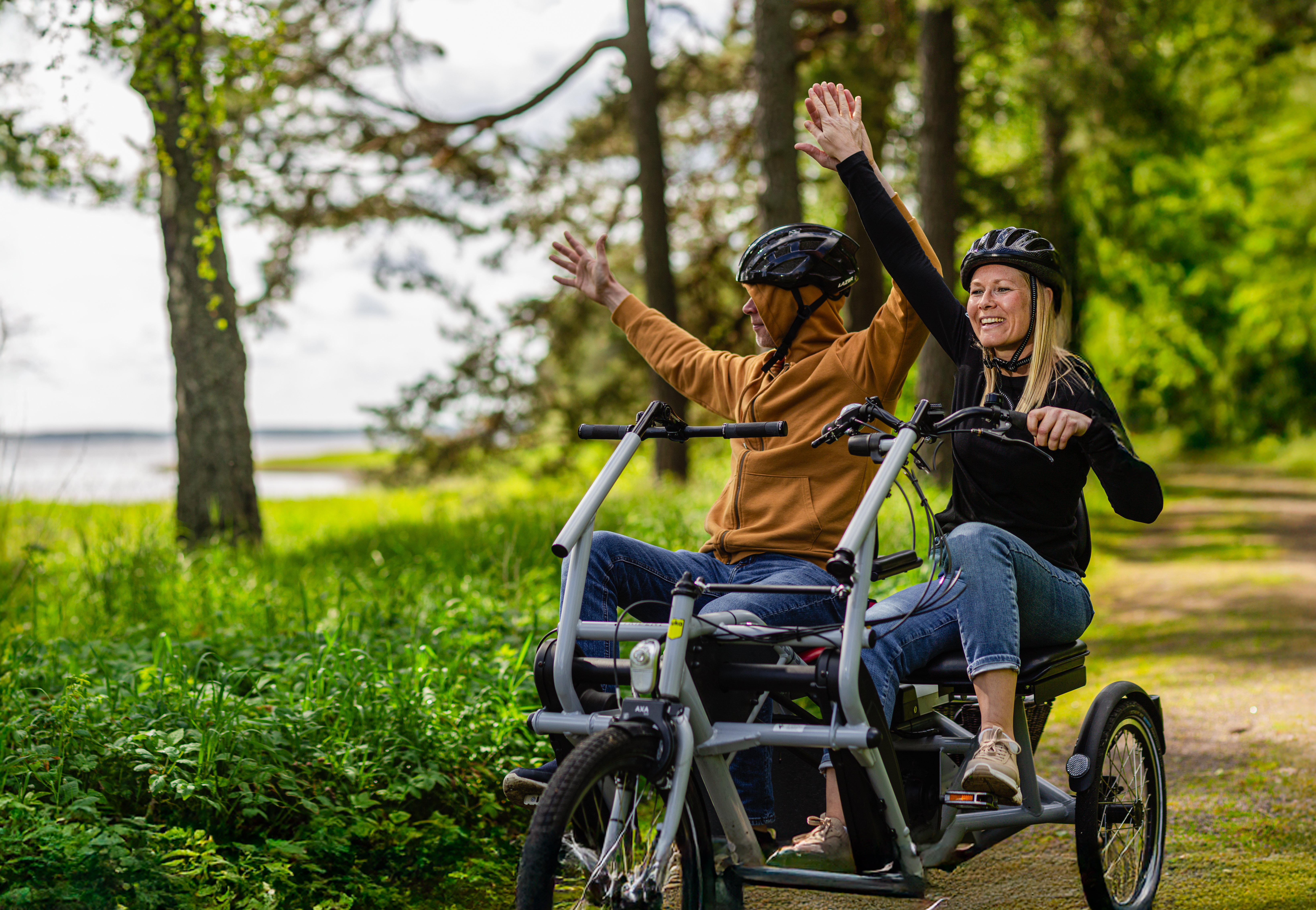 Två personer med cykelhjälmar sitter tillsammans på en trehjulig cykel på en skogsstig. De håller upp händerna och ser glada ut medan de cyklar genom det gröna landskapet.