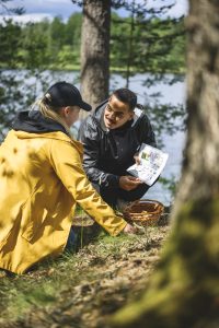 Två personer sitter på marken i en skog nära en sjö. Den ena håller i ett papper med bilder och symboler, medan den andra plockar något från marken och lägger i en korg. 